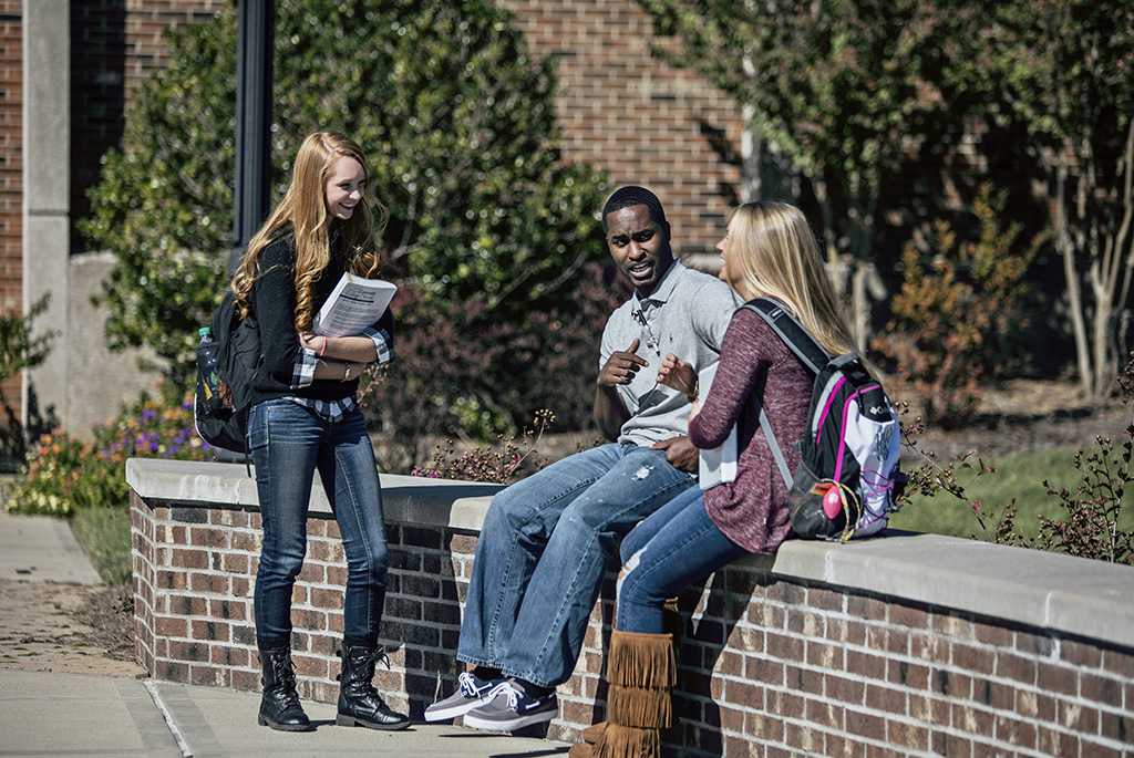 One female student standing talking to a male and female student sitting on a brick wall on campus