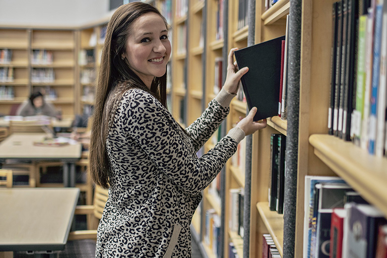 Woman smiling at camera while pulling book out of library bookshelf