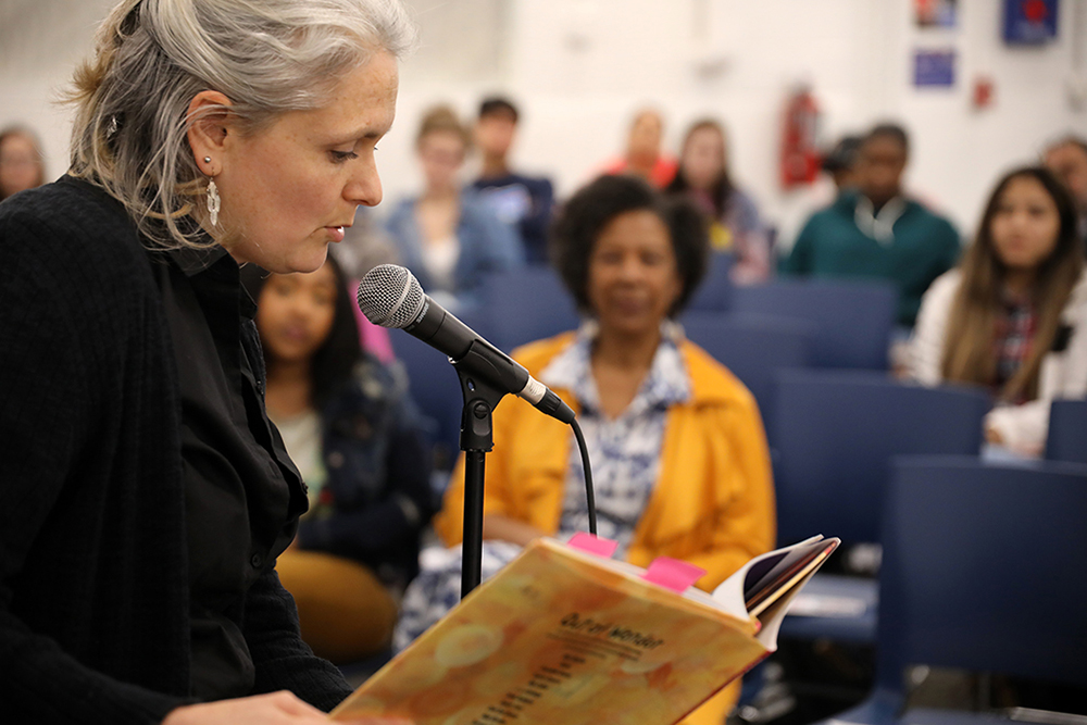 Lady reading a book at microphone with audiance in the background