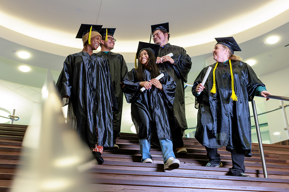 Five graduates dressed in cap and gown walking down stairs