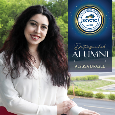 Alyssa Brasel standing in the courtyard at the college with banner that says Distinguished Alumni with blue banner and college crest