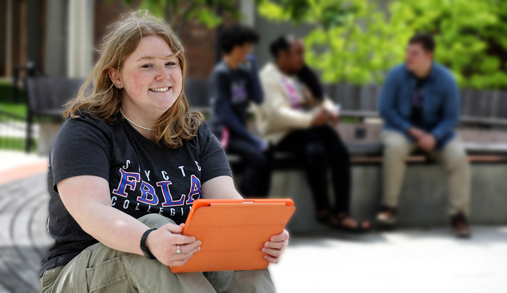 Ann Riley setting on a bench holding her IPad with student in the background