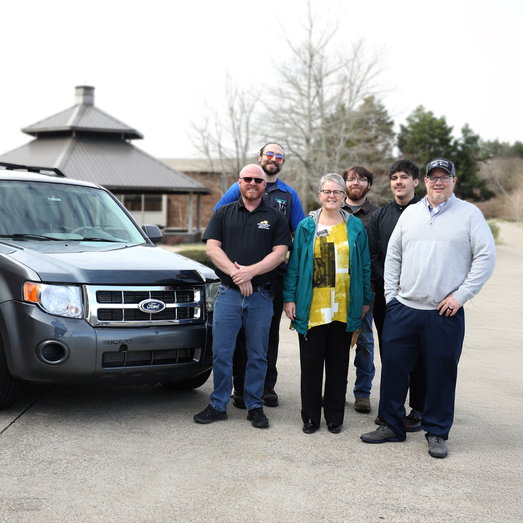 A group of six people stand next to a donated automobile.