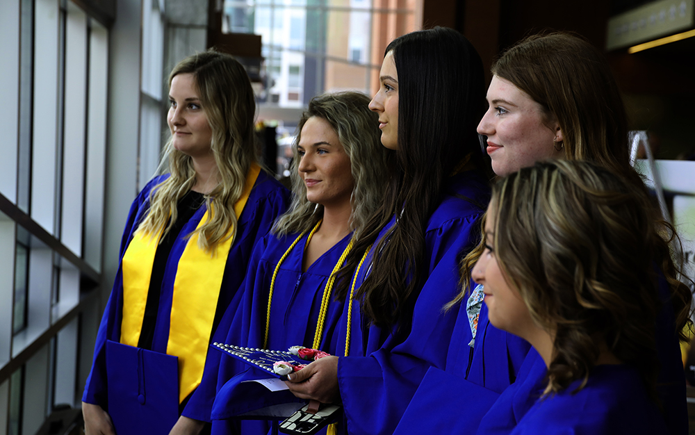 Five ladies in cap and gown looking out the window at graduation.