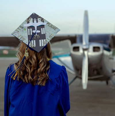 Photo of Mary Schalk facing an airplane with her graduation cap and gown on.