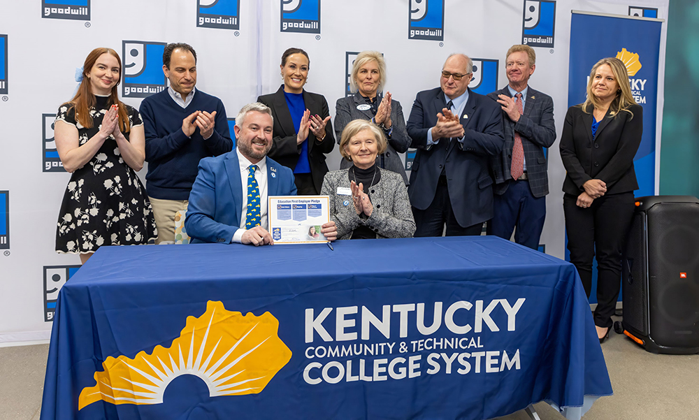 Two people seated holding up teh Education First Employer sign with six people behind clapping