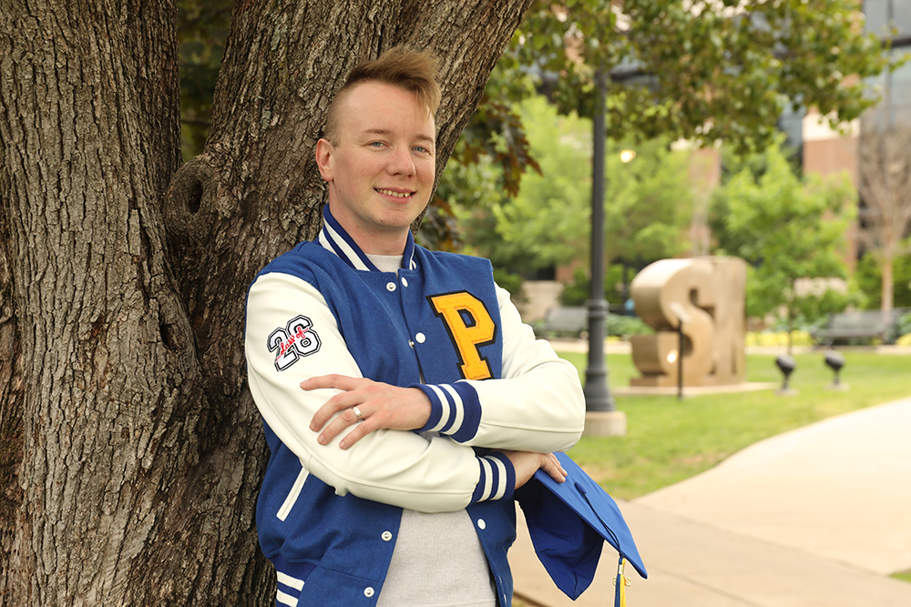 Alex Smith with arms crossed and Pathfinder Letterman jacket on leaning on a tree on campus
