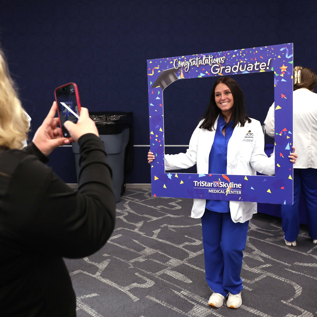 Female Allied Health Student wearing scrubs holding a photo frame with someone taking her photo.