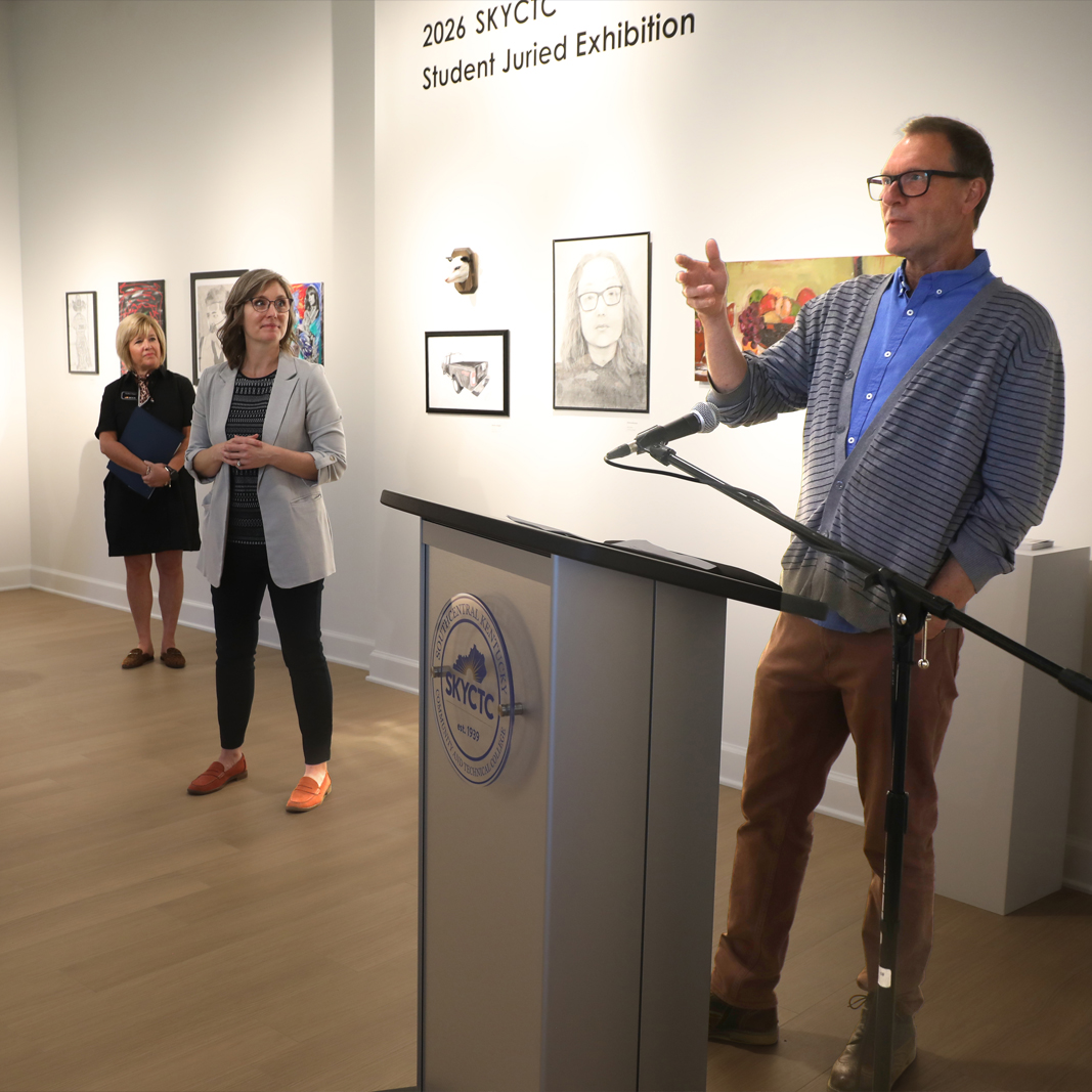 Man speaking at podium with two ladies listing in background