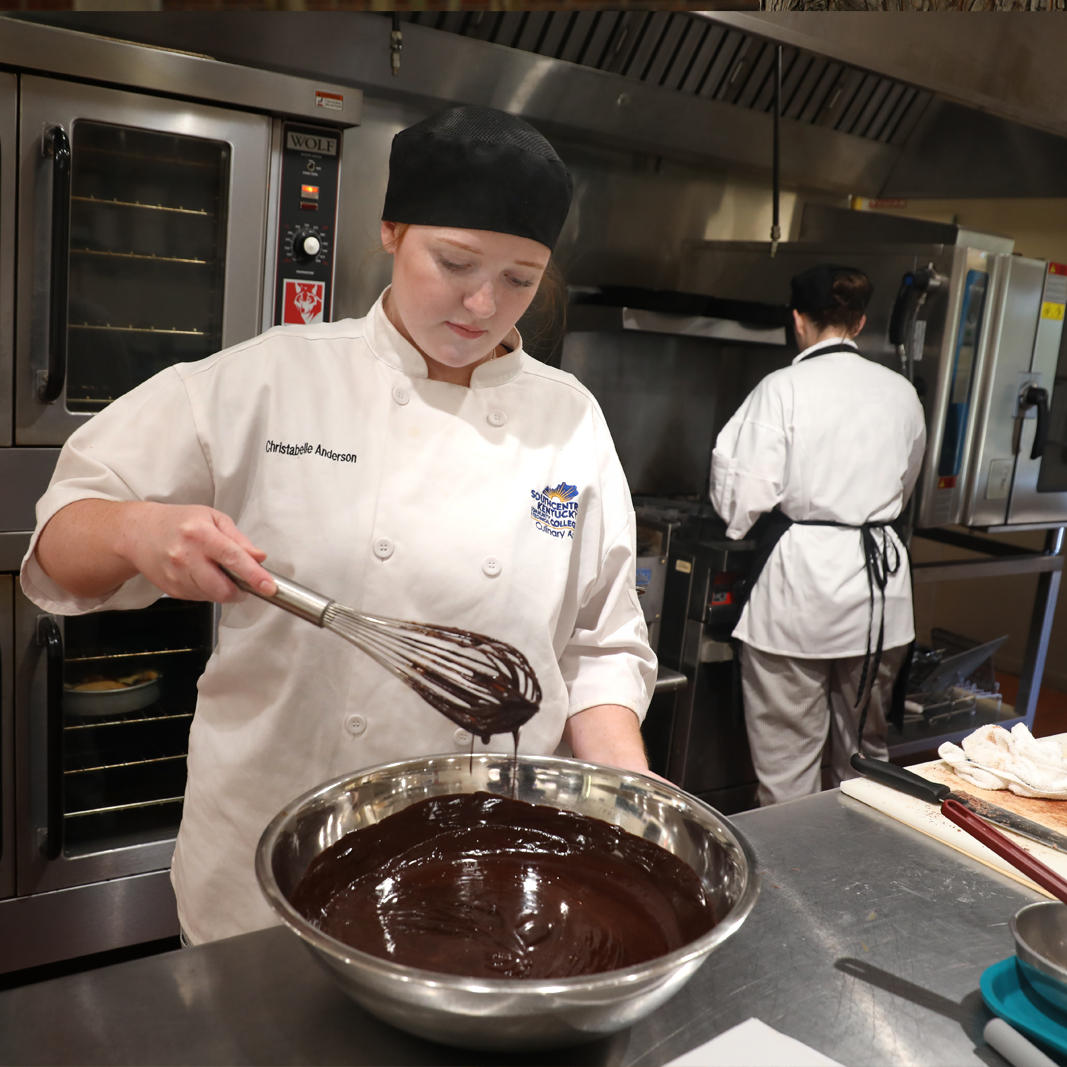 Christabelle Anderson with chef uniform on in a kitchen stirring a large bowl of chocolate.