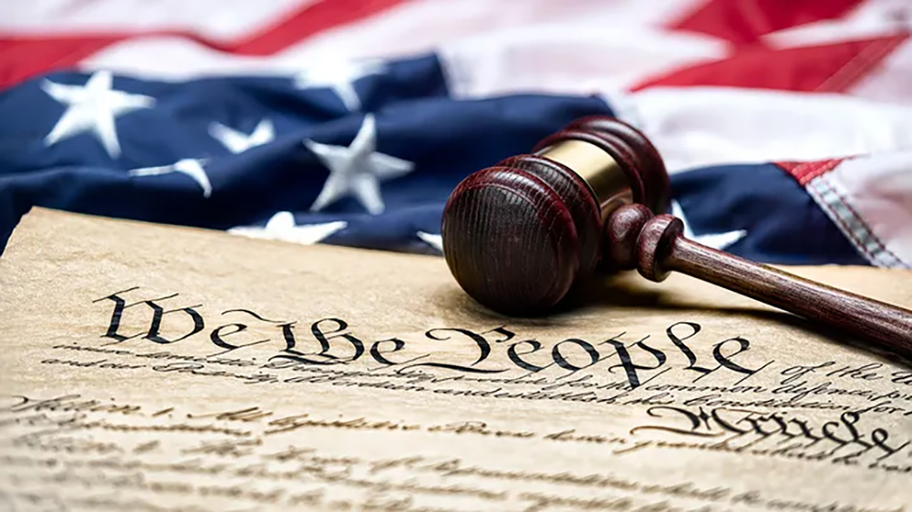 Image of a copy of the original U.S. Constitution with a gavil laying on it and thee American Flag is the background