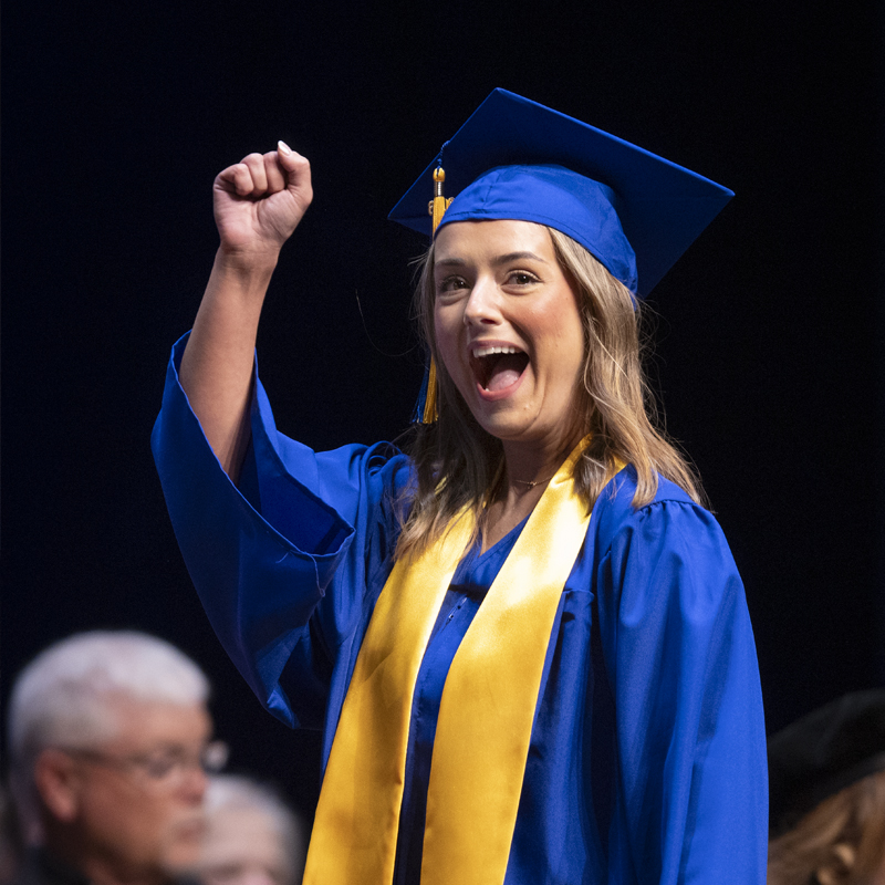 Dual Credit student walking across stage in cap and gown with had raised.