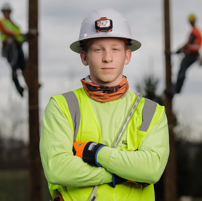 A young lineman dressed in hard ad and uniform with his hands crossed and two linemen on poles in the background
